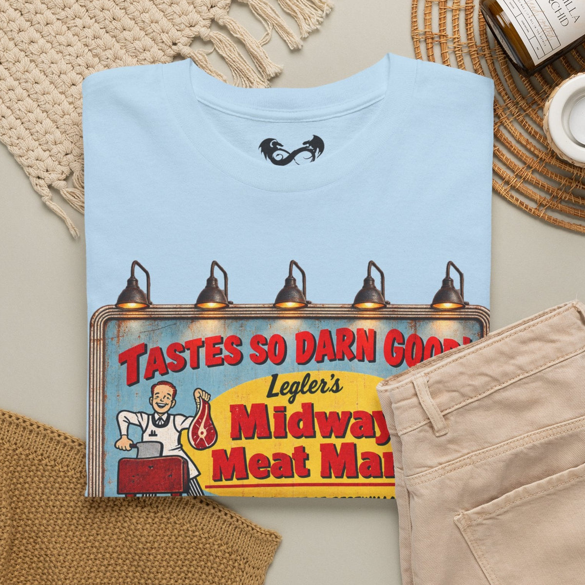 A folded light blue T-shirt with a colorful retro meat market sign graphic on the front, styled in a flat lay with beige pants, a knit blanket, a woven mat, and a small candle jar on a light neutral background.