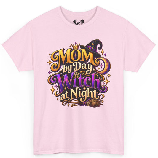 A light pink short-sleeve crewneck t-shirt featuring a colorful graphic on the front that reads 'MOM by Day, Witch at Night' in stylized gold, purple, and black lettering with a witch hat and broomstick illustration.