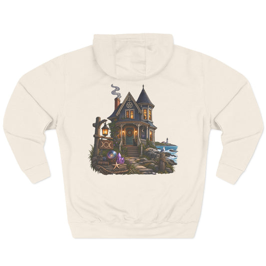 An off-white hooded sweatshirt shown from the back, featuring a detailed graphic print of a haunted house with a lantern, a small boat, and a purple creature on a stone path.