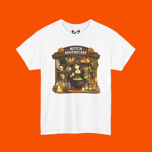 A white short-sleeve crewneck t-shirt featuring a colorful graphic print of a witch apothecary shop with pumpkins, cauldron, and potion bottles.