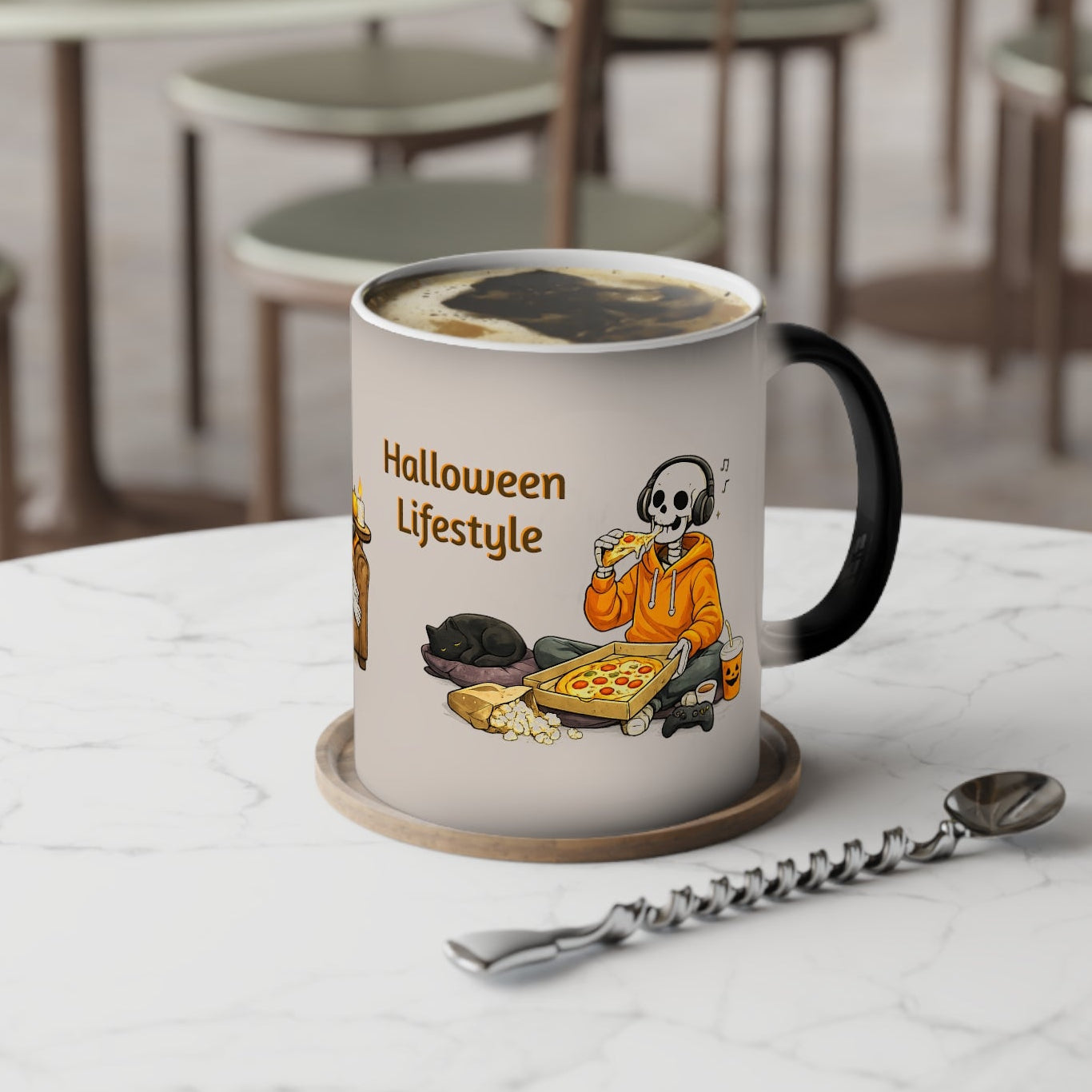 A white ceramic coffee mug with a black handle sits on a marble table in a cafe setting. The mug features a cartoon graphic of a skeleton in an orange hoodie eating pizza with the text 'Halloween Lifestyle'.