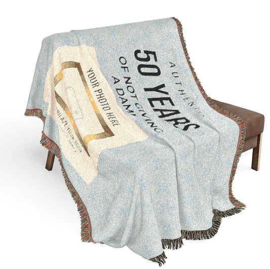 A woven throw blanket draped over a brown bench against a white studio background. The blanket has a light blue and white speckled base with a large cream graphic area and black text reading '50 YEARS' and 'AUTHENTIC'.