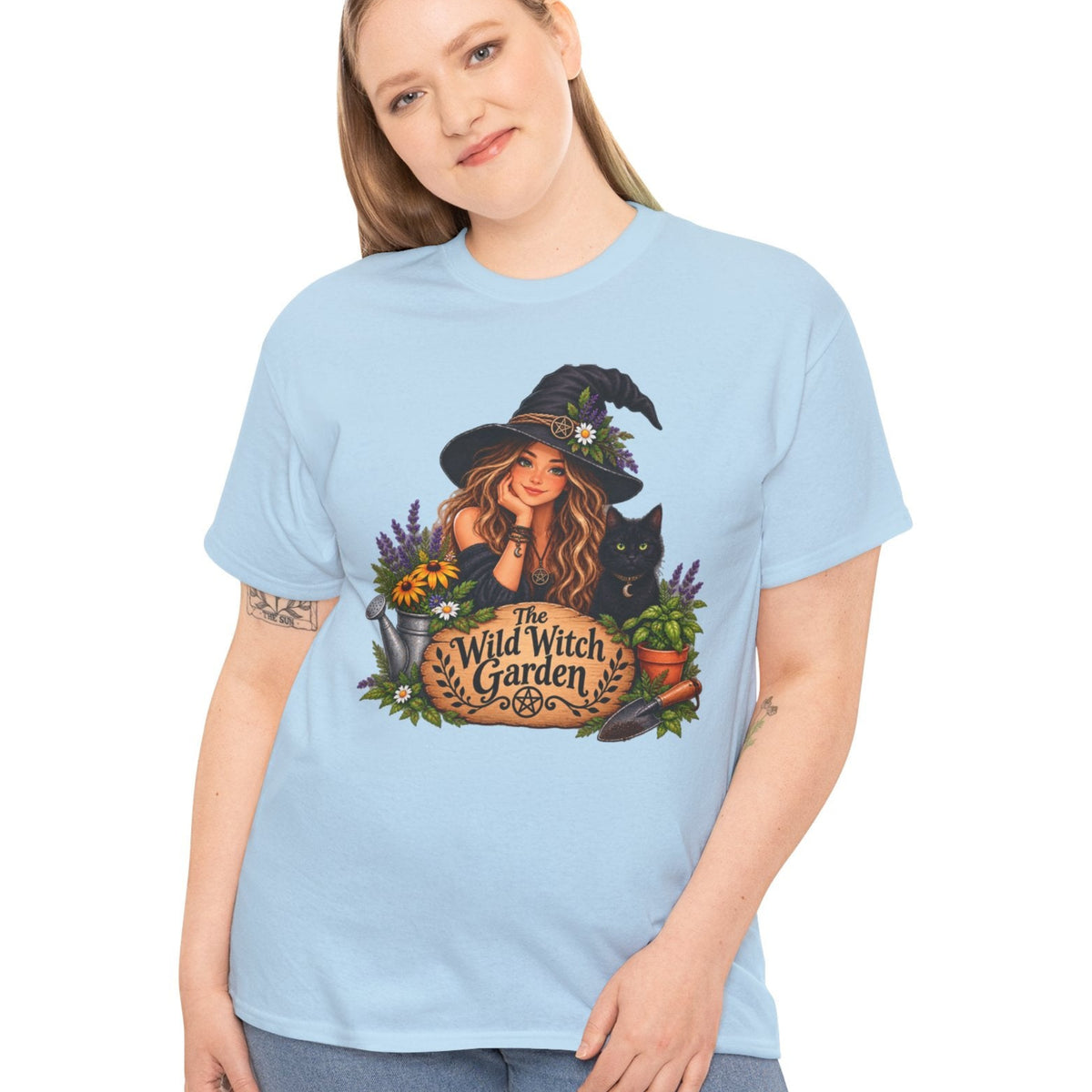 A woman modeling a light blue crewneck t-shirt with a colorful graphic of a witch in a hat next to a sign that reads 'The Wild Witch Garden'.