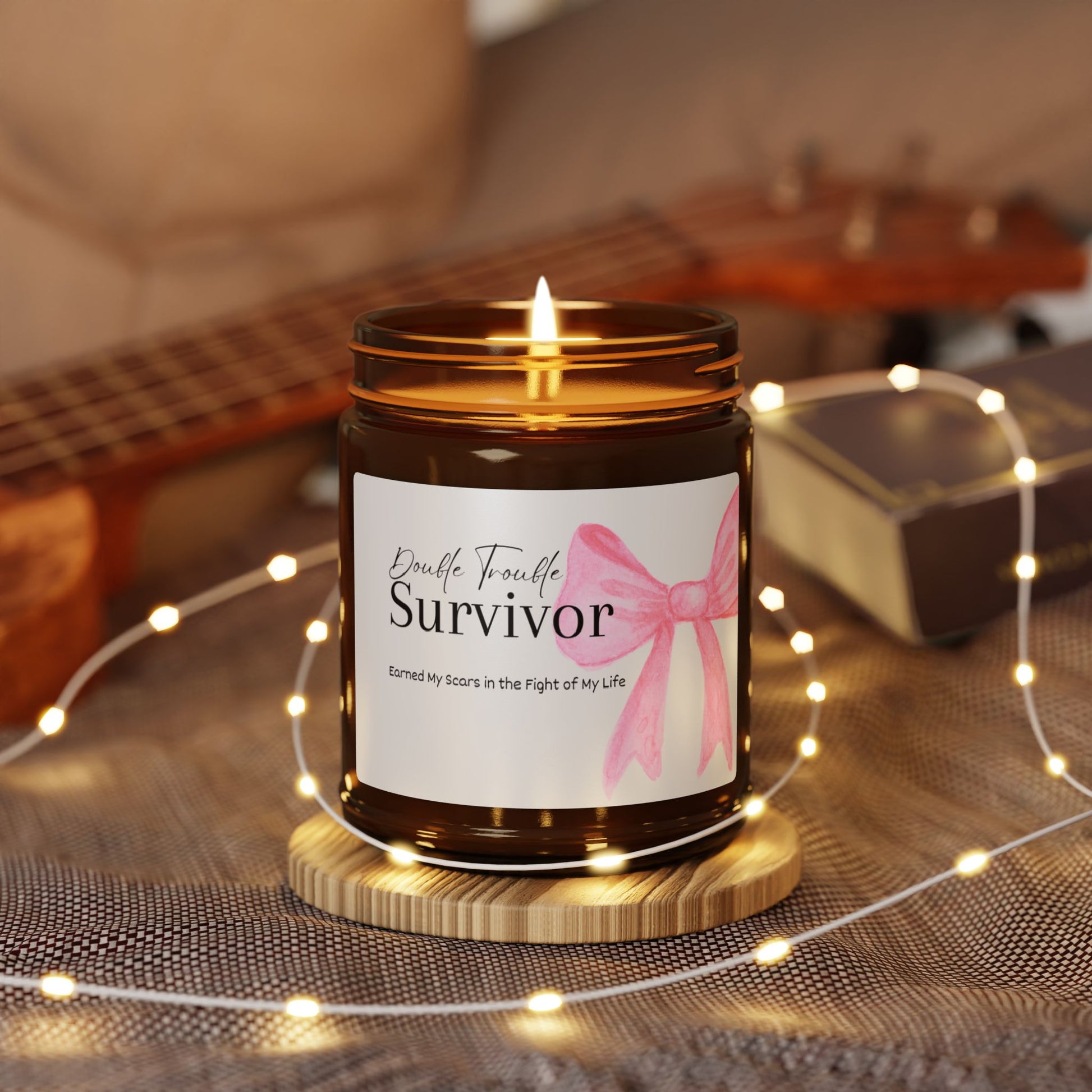 Candle in a jar labeled 'Survivor' with a pink ribbon on a wooden coaster, surrounded by string lights and a guitar in the background.
