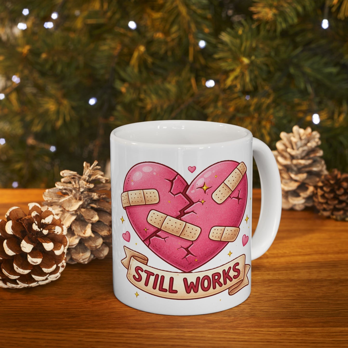 White mug with a heart design and 'STILL WORKS' text, placed on a wooden surface with pinecones and a Christmas tree in the background.