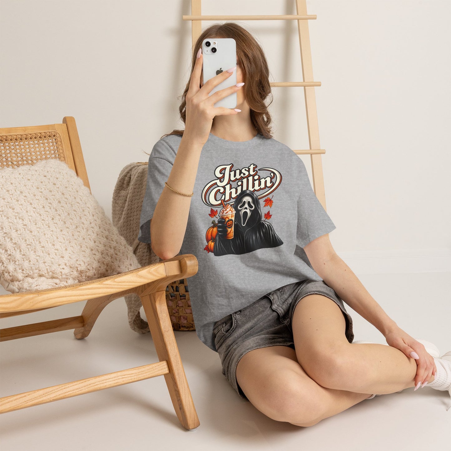 A woman takes a mirror selfie while wearing a heather gray graphic t-shirt with a 'Just Chillin'' Ghostface design, paired with denim shorts and white sneakers, sitting on a wooden chair in a minimalist room.
