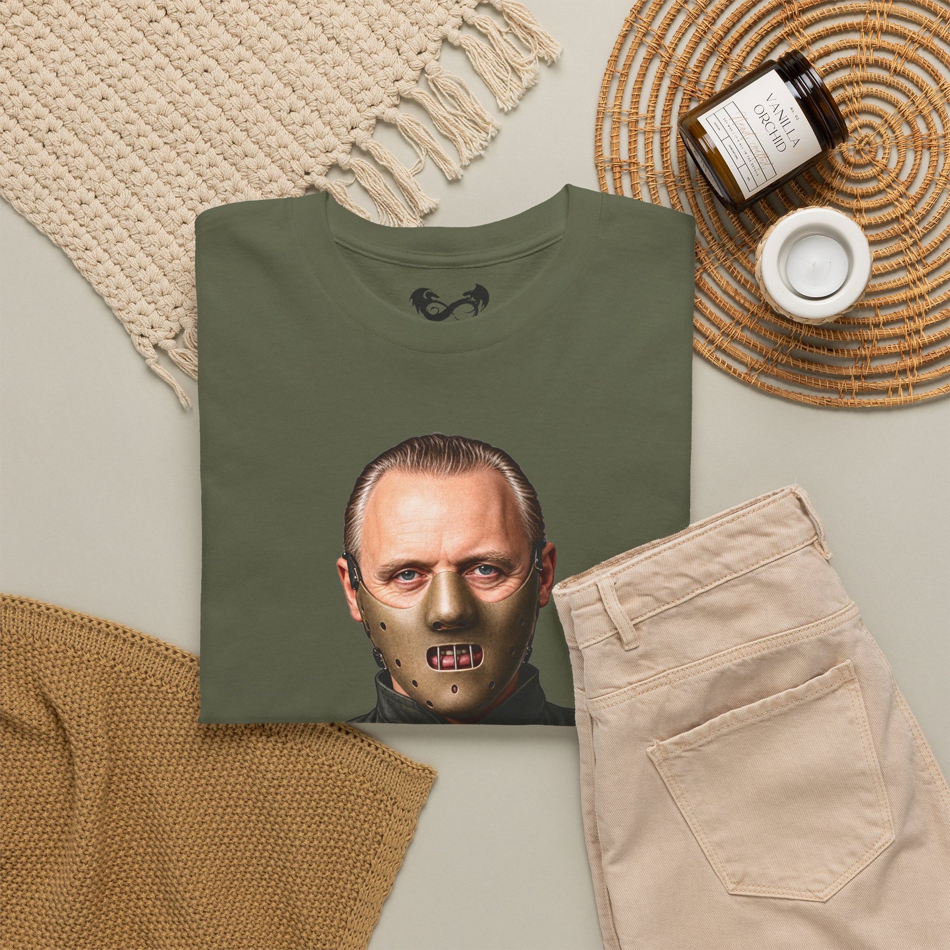 An olive green t-shirt featuring a graphic print of Jason Voorhees' face with a metallic hockey mask, styled in a flat lay with beige pants, a knit blanket, a candle, and a jar on a neutral background.