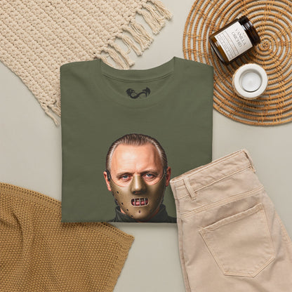An olive green t-shirt featuring a graphic print of Jason Voorhees' face with a metallic hockey mask, styled in a flat lay with beige pants, a knit blanket, a candle, and a jar on a neutral background.