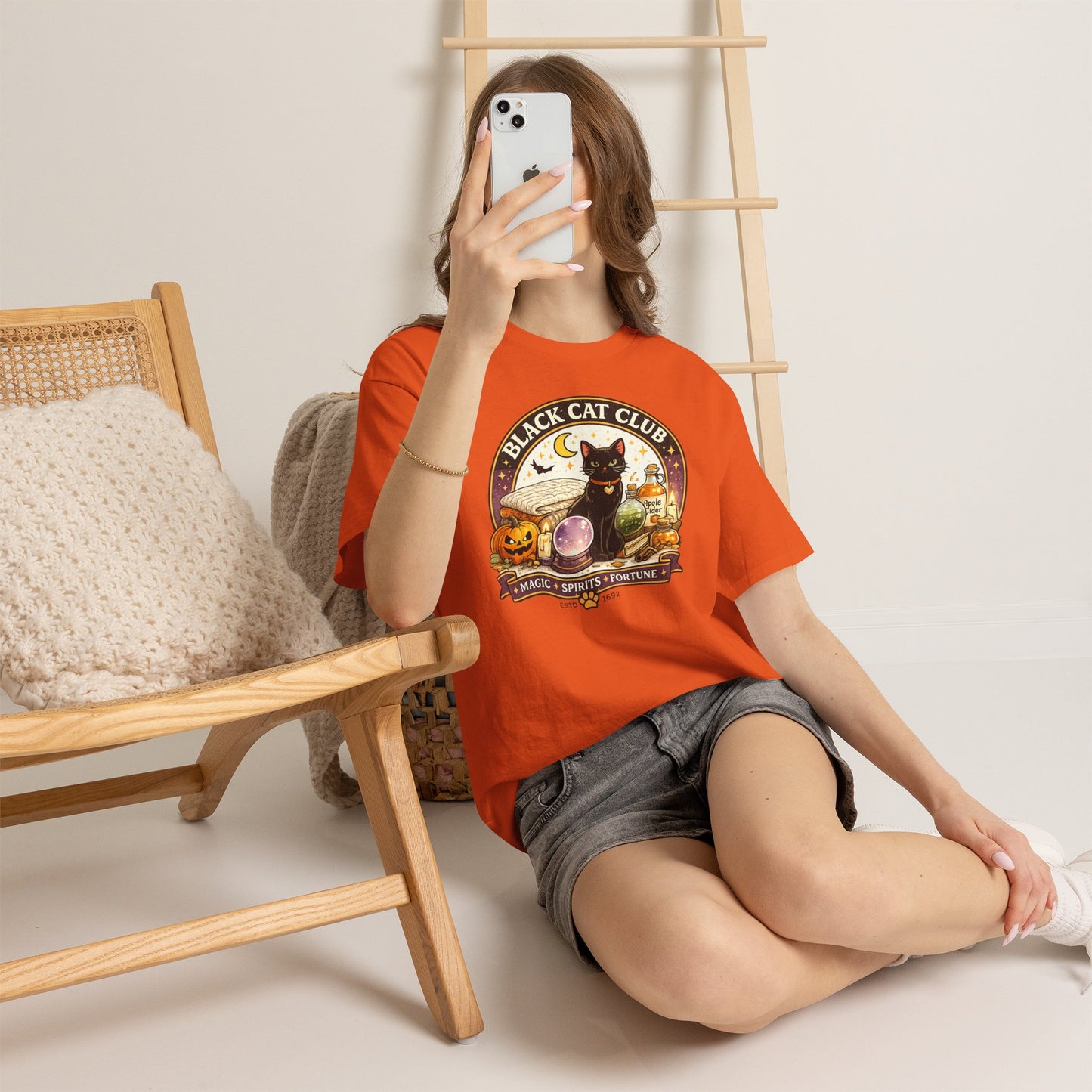 A woman takes a mirror selfie while wearing a bright orange graphic t-shirt with a 'Black Cat Club' design, paired with denim shorts, in a cozy indoor setting with a wooden chair and ladder.