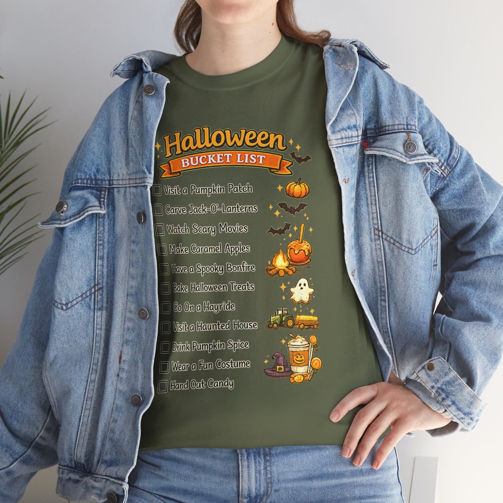 A person wearing an olive green graphic T-shirt with a Halloween bucket list design, layered under a light-wash denim jacket. The graphic features orange pumpkins, bats, a ghost, and a checklist of spooky activities.