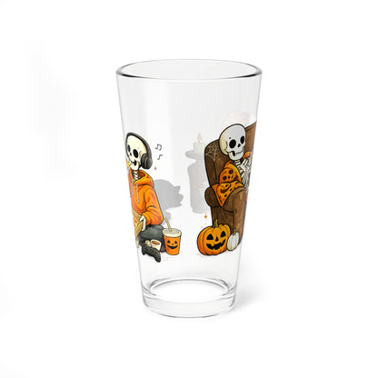 A clear pint glass featuring a cartoon Halloween illustration of two skeleton characters, one smoking a pipe and the other lounging on a couch with a pumpkin, shown on a white background.