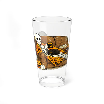 A clear drinking glass featuring a Halloween-themed illustration of a skeleton lounging on a couch with pizza, popcorn, and a black cat, shown on a white background.
