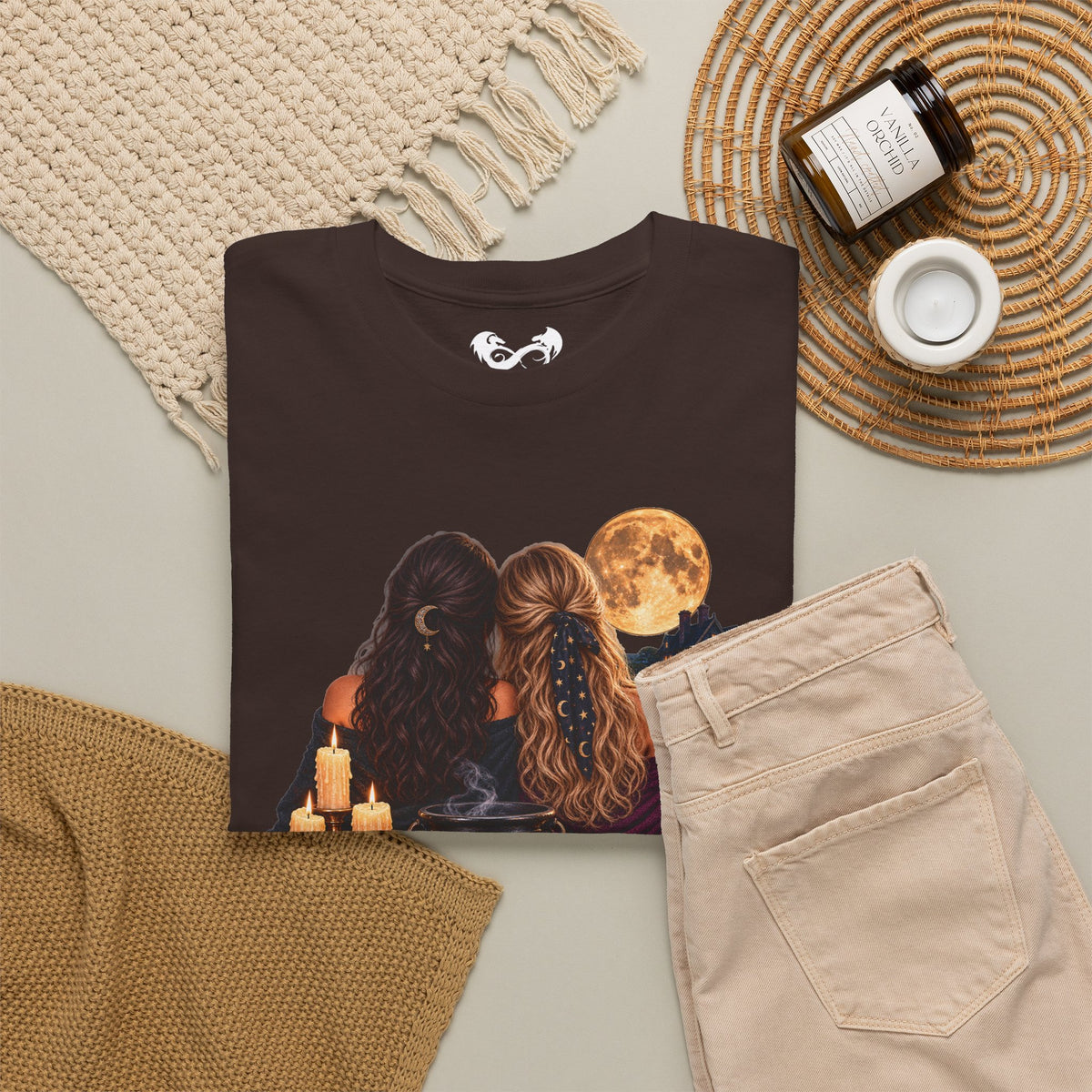 A black graphic t-shirt featuring an illustration of two women with long hair facing a full moon, styled in a cozy flat-lay with beige pants, a knit blanket, a candle, and a jar on a neutral background.