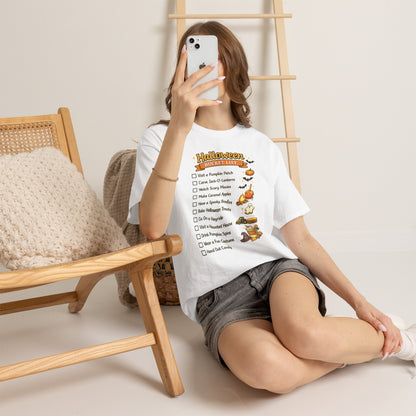 A woman sits on a floor in a cozy room setting, wearing a white graphic t-shirt with a 'Halloween Chores' checklist and gray denim shorts. She is holding a white smartphone to take a mirror selfie, with a wooden chair and ladder in the background.