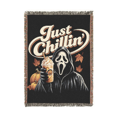 A rectangular woven throw blanket with fringe on all sides, featuring a black background and a graphic of the Ghostface character holding a drink, with the phrase 'Just Chillin'' in large cream script and autumn leaves and pumpkins around him.