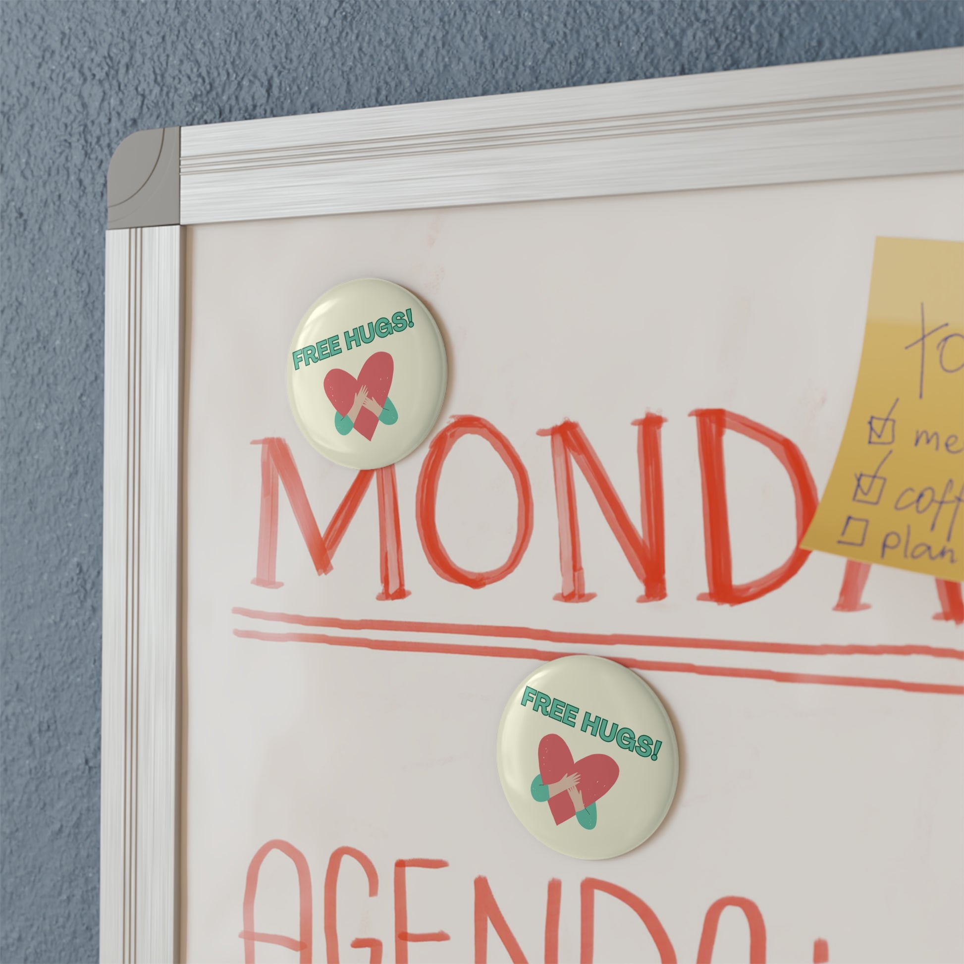 Whiteboard with 'MONDAY' and 'AGENDA' text, buttons with 'FREE HUGS!' text, and a yellow sticky note on a blue wall.