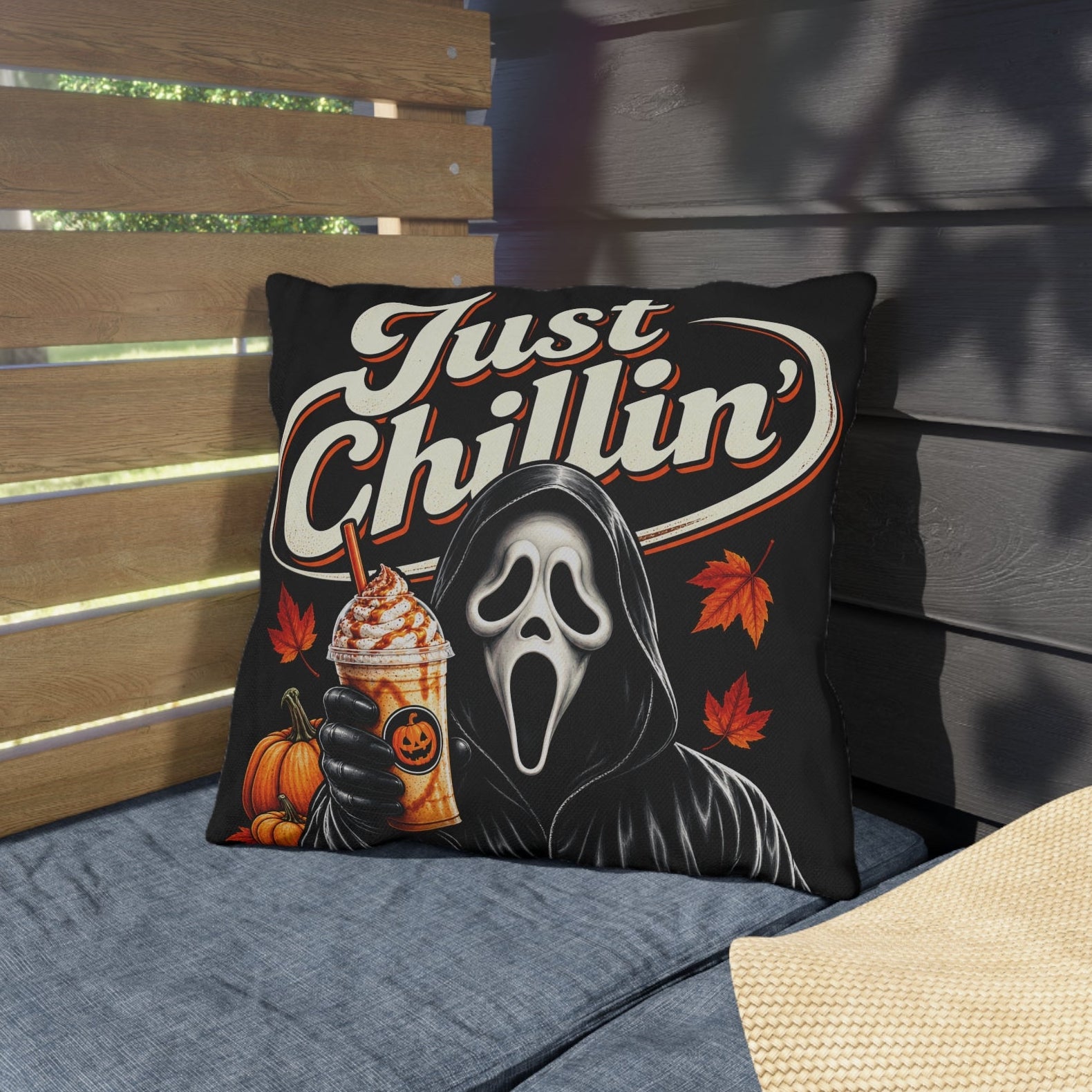 A square throw pillow with a Ghostface 'Just Chillin'' graphic, styled on a blue outdoor patio cushion next to a straw hat.