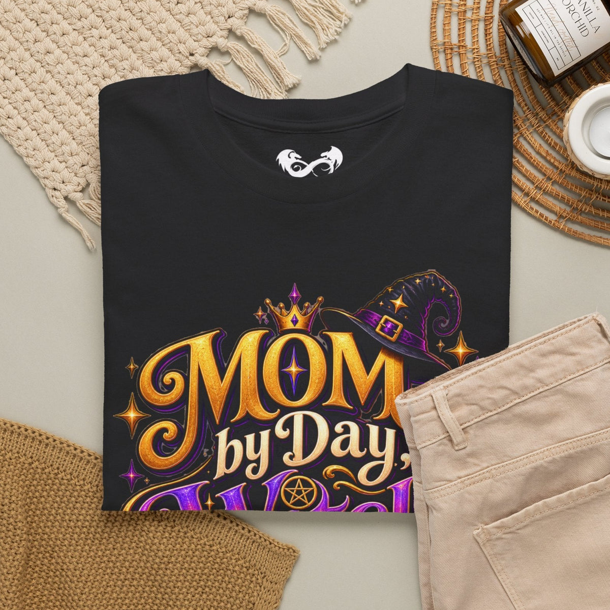 A folded black graphic T-shirt is arranged in a styled flat lay with beige pants, a woven rug, a knit blanket, a candle jar, and a small cup on a neutral surface. The shirt graphic reads “MOM by Day” in large gold lettering with purple and gold decorative elements.