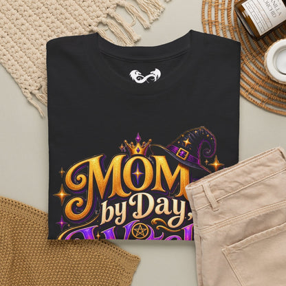 A folded black graphic T-shirt is arranged in a styled flat lay with beige pants, a woven rug, a knit blanket, a candle jar, and a small cup on a neutral surface. The shirt graphic reads “MOM by Day” in large gold lettering with purple and gold decorative elements.