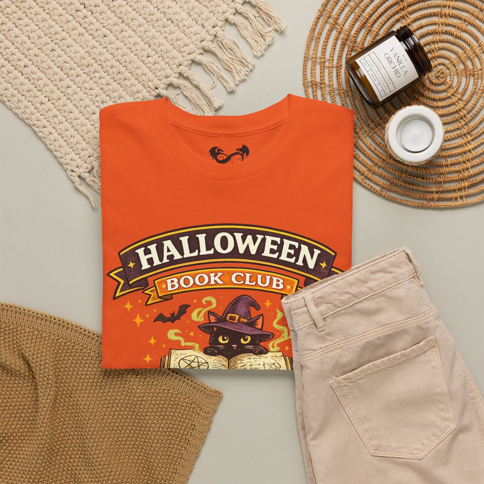 An orange graphic T-shirt laid flat on a light gray surface, styled with a cream knit blanket, tan knit fabric, a woven placemat, a candle jar, and a small white cup. The shirt reads "HALLOWEEN BOOK CLUB" with a witch cat illustration and stars on the front.
