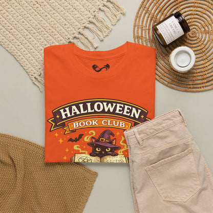 An orange graphic T-shirt laid flat on a light gray surface, styled with a cream knit blanket, tan knit fabric, a woven placemat, a candle jar, and a small white cup. The shirt reads "HALLOWEEN BOOK CLUB" with a witch cat illustration and stars on the front.