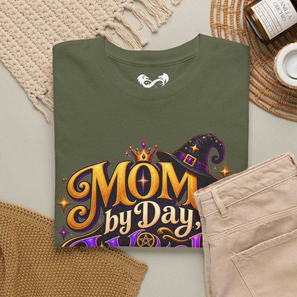 An olive green t-shirt with a 'MOM by Day' witch graphic, styled in a flat lay with beige pants, a knit blanket, a candle, and a cup on a neutral background.