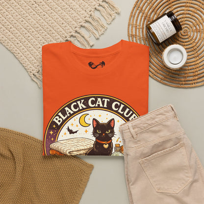 An orange folded graphic T-shirt is styled in a flat lay with beige pants, a cream knit blanket, a woven mat, a candle jar, and a small white cup on a light neutral background. The shirt graphic reads 'BLACK CAT CLUB' with an illustration of a black cat, moon, stars, and bats.