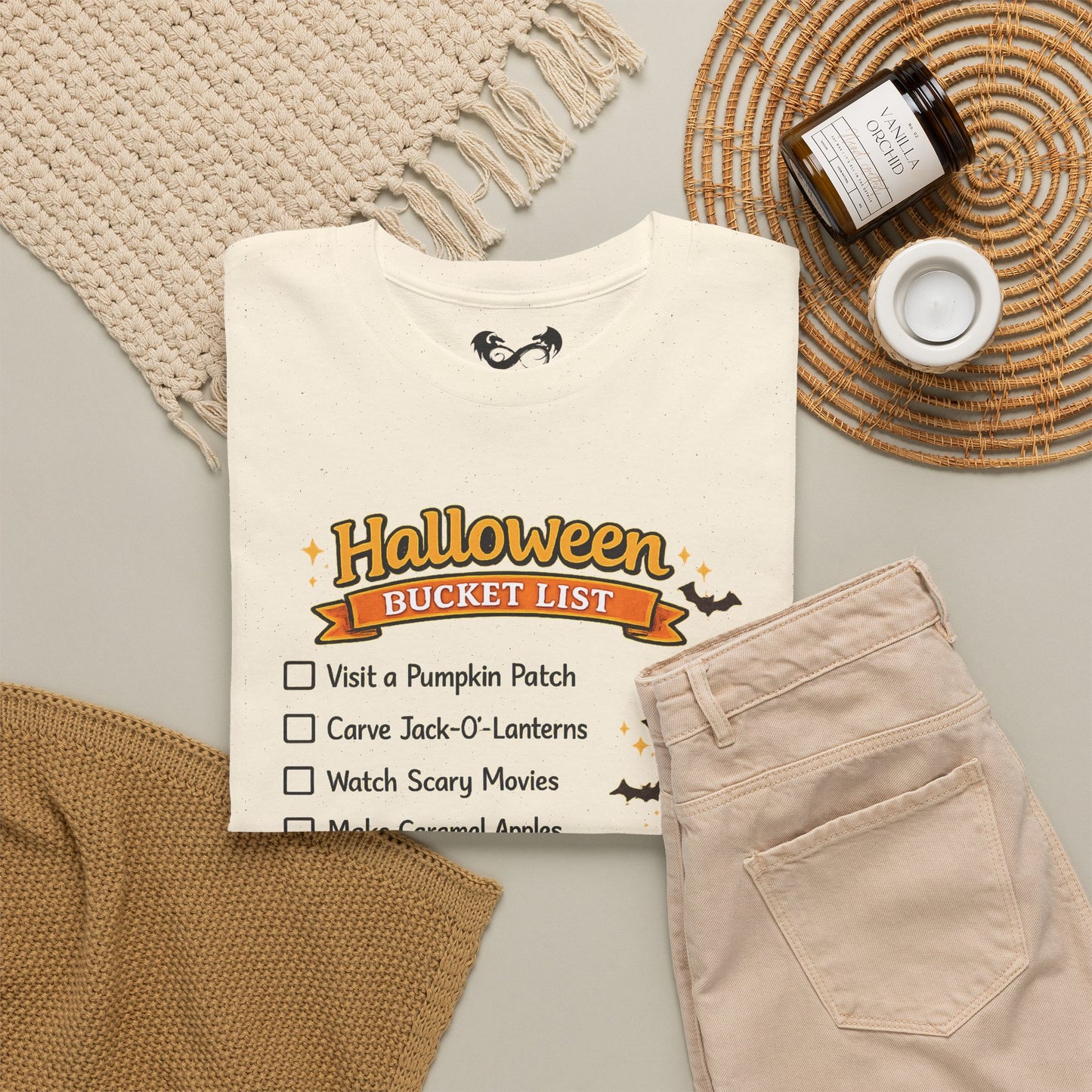An off-white t-shirt with a 'Halloween Bucket List' graphic, styled in a flat lay with beige pants, a knit blanket, a jar candle, and a woven placemat on a light gray background.