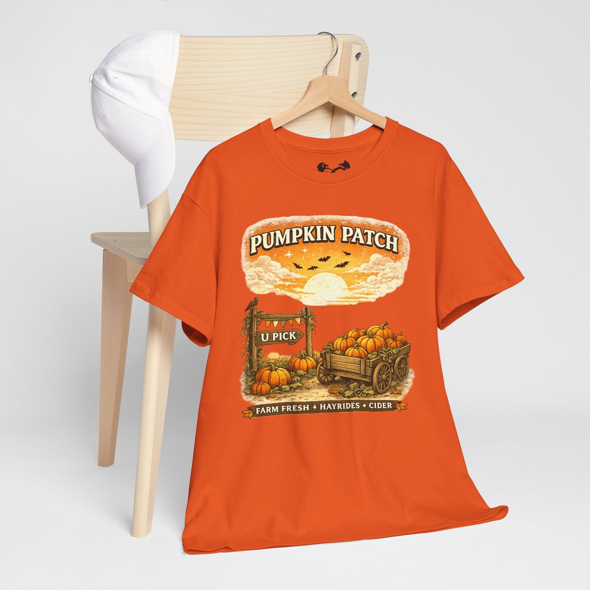 An orange graphic t-shirt hanging on a wooden hanger from a light wood chair. The t-shirt features a 'Pumpkin Patch U Pick' autumn graphic with pumpkins, a wagon, and a sunset.
