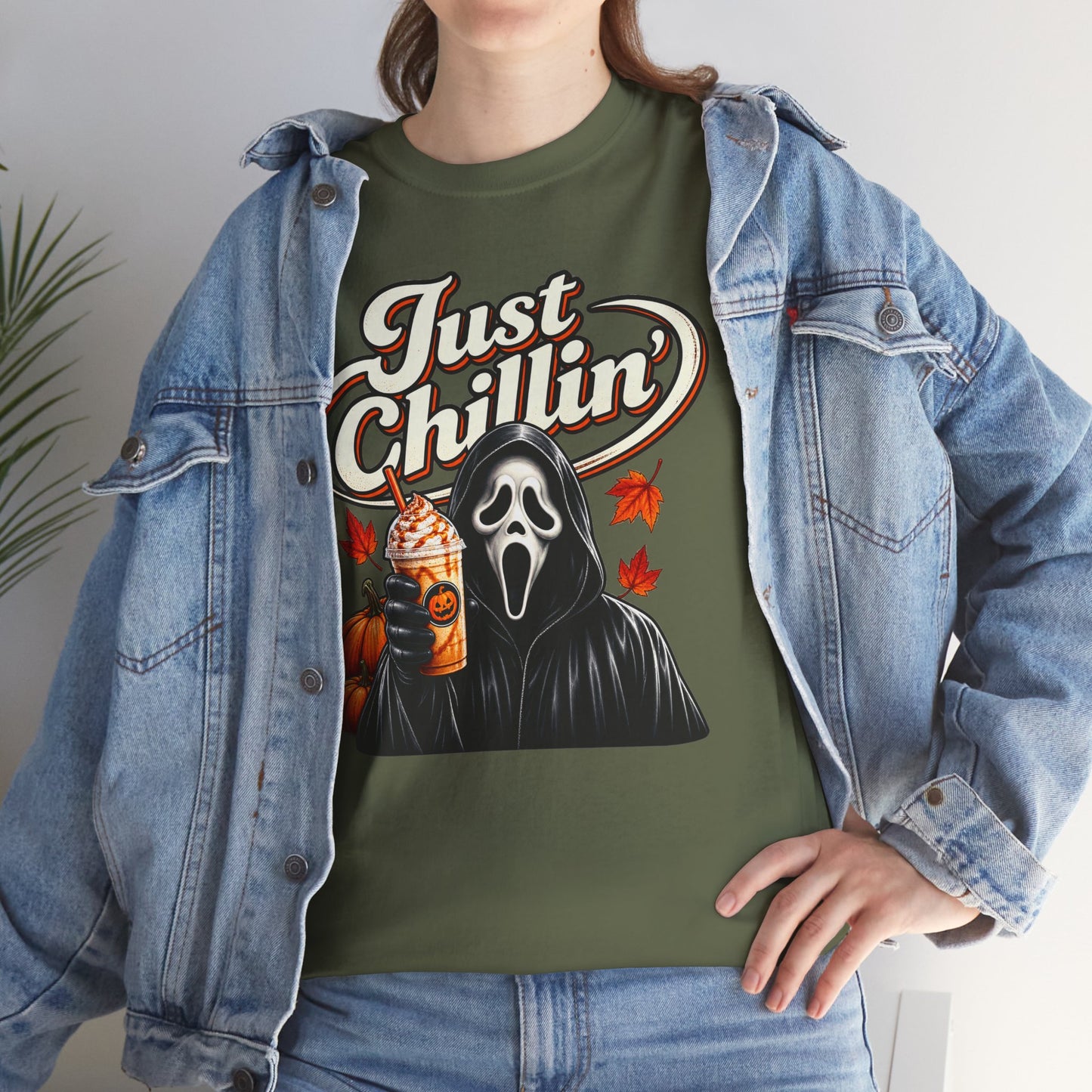 A person wearing an olive green graphic t-shirt under an open light-wash denim jacket. The t-shirt features a Ghostface-style hooded figure holding an orange drink with the text 'Just Chillin'' in white script.
