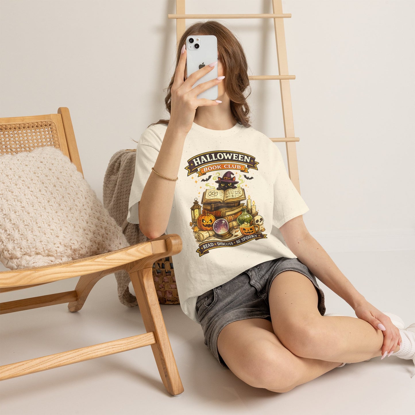 A woman takes a mirror selfie while wearing a cream-colored graphic t-shirt that reads 'HALLOWEEN SPooktacular' with a spooky pumpkin and coffin illustration, paired with denim shorts in a cozy indoor setting.