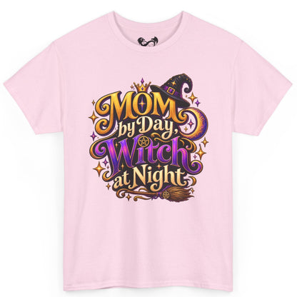 A light pink short-sleeve crewneck t-shirt featuring a colorful graphic on the front that reads 'MOM by Day, Witch at Night' in stylized gold, purple, and black lettering with a witch hat and broomstick illustration.