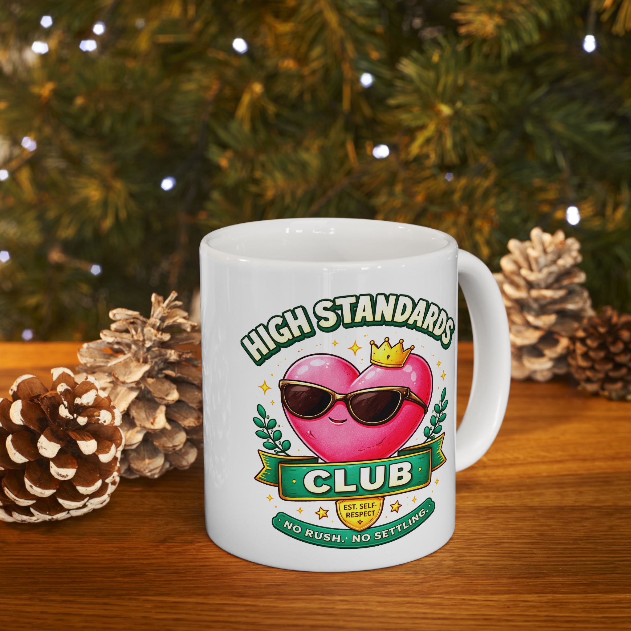 White mug with a heart design and text 'High Standards Club' on a wooden surface with pinecones and a Christmas tree in the background.