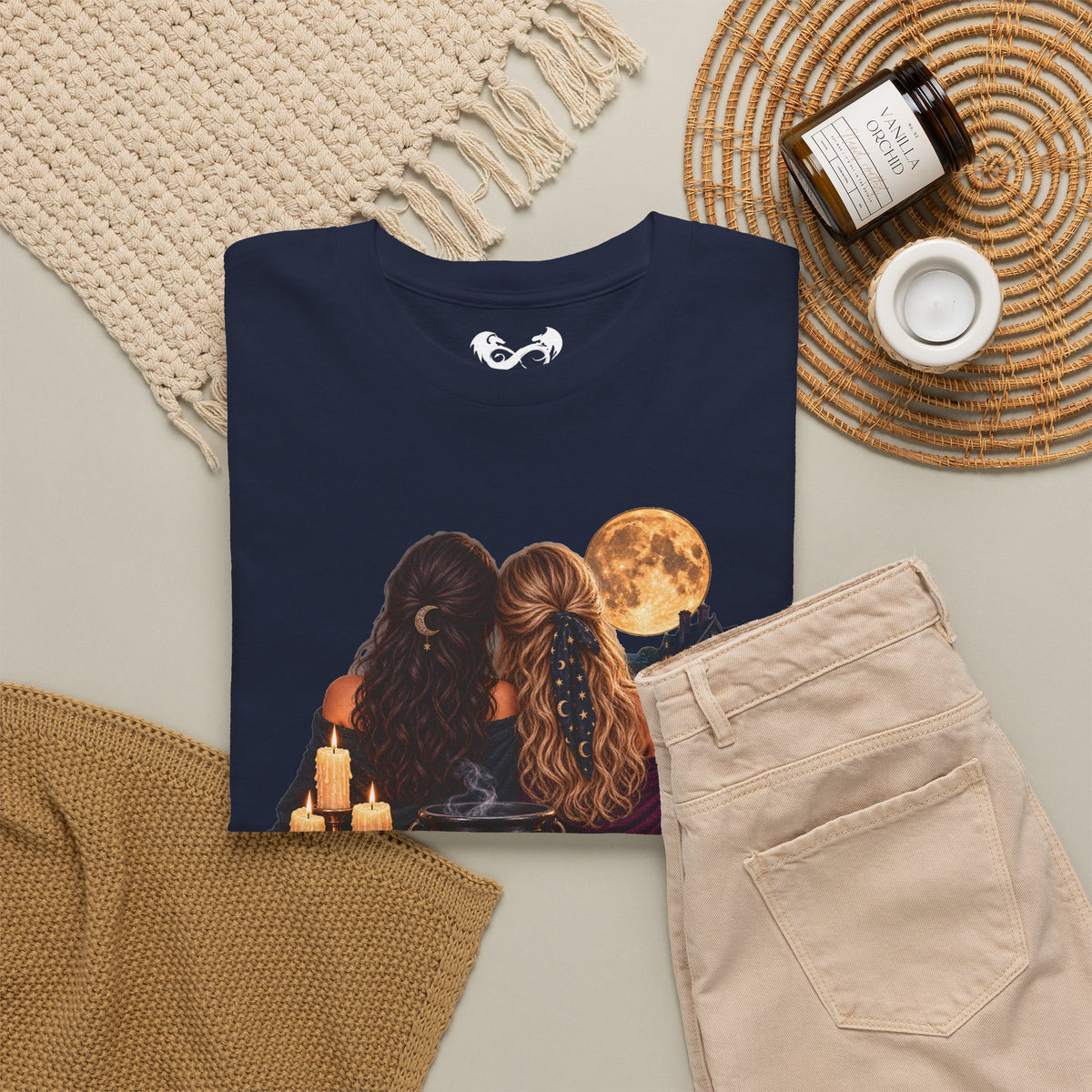 A black graphic t-shirt featuring an illustration of two women with long hair facing a full moon, styled in a cozy flat-lay with beige pants, a knit blanket, a candle, and a jar on a neutral background.