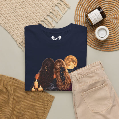 A black graphic t-shirt featuring an illustration of two women with long hair facing a full moon, styled in a cozy flat-lay with beige pants, a knit blanket, a candle, and a jar on a neutral background.