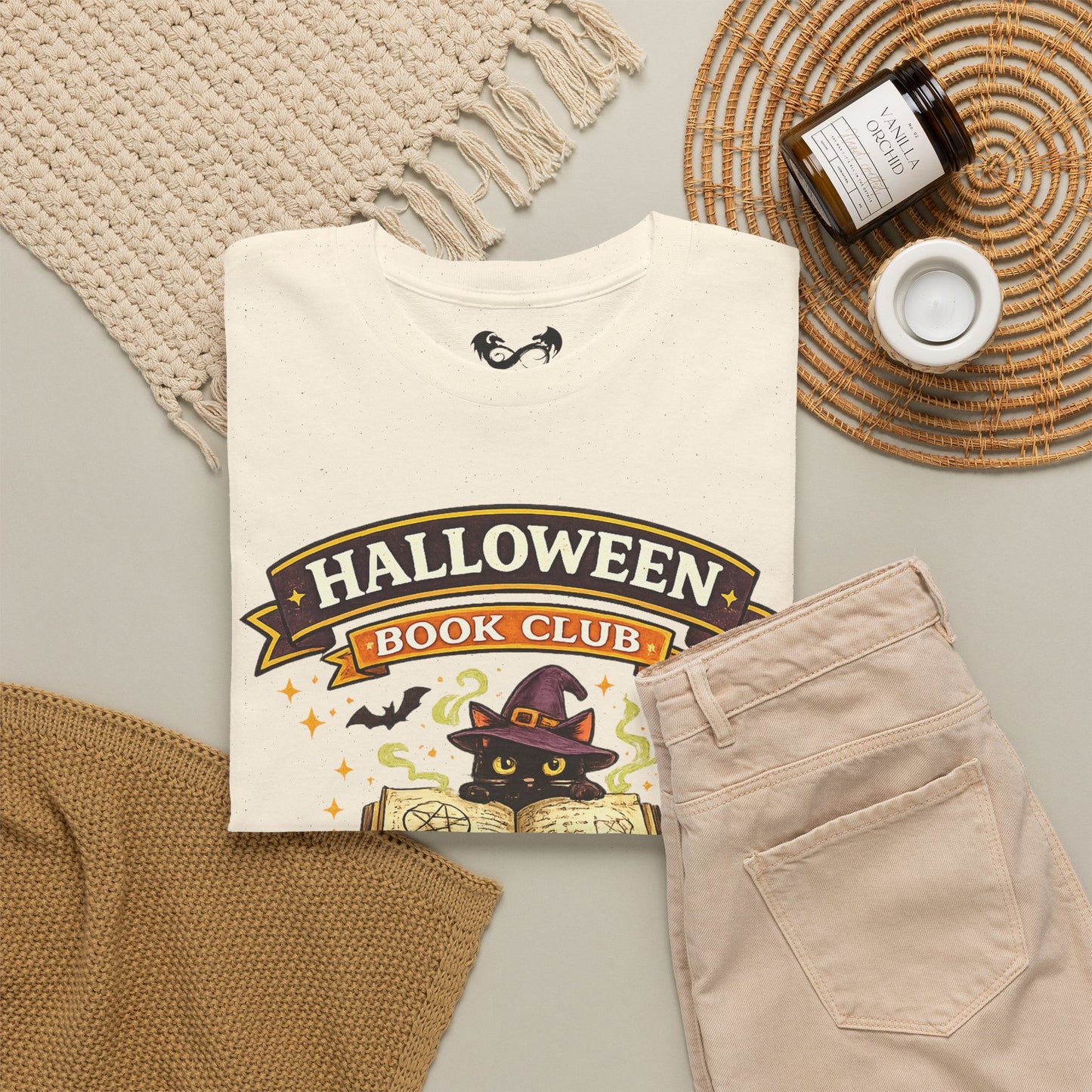 A folded cream graphic T-shirt with a Halloween-themed book club print, styled in a flat lay with beige pants, a woven blanket, a small candle, and a jar on a light gray surface.