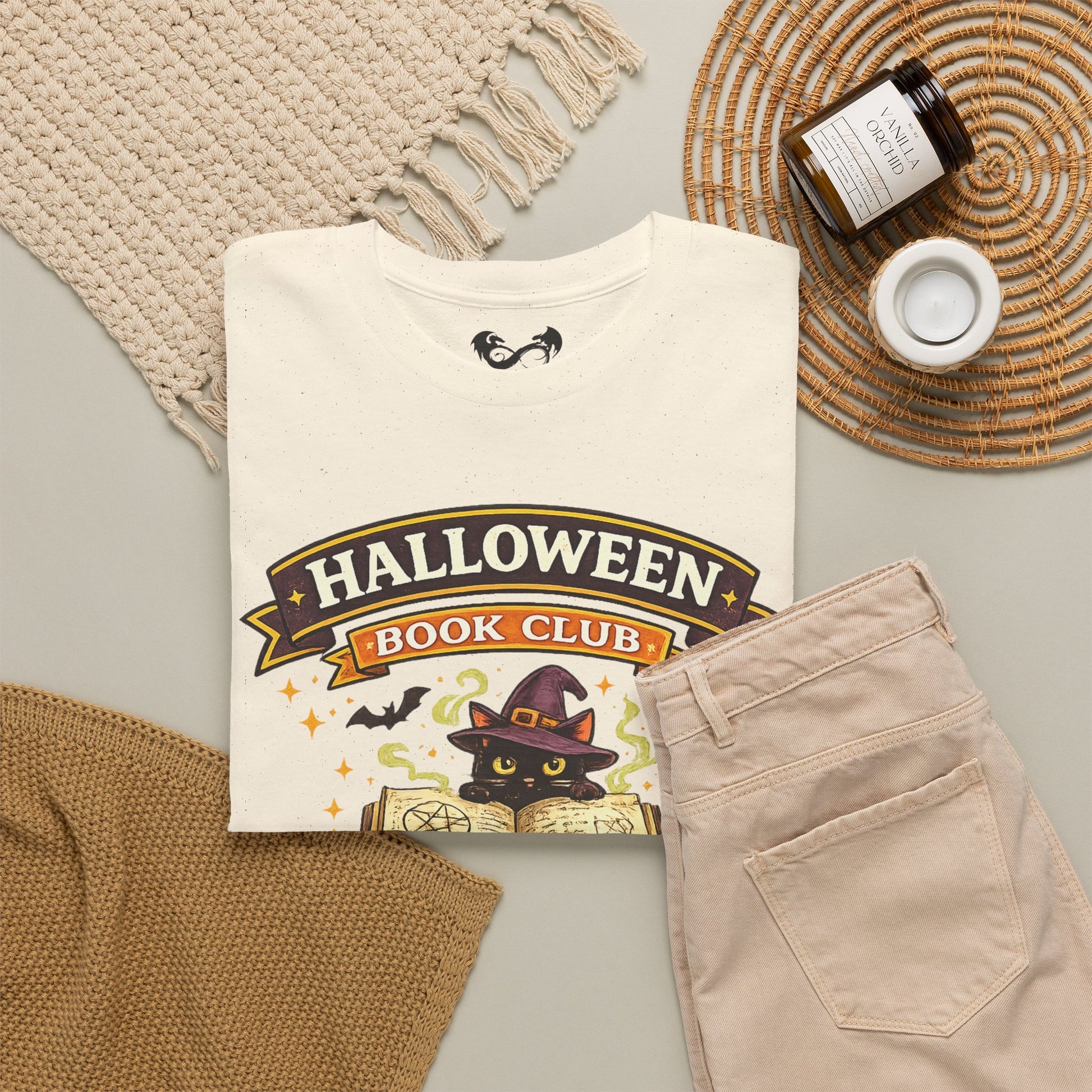 A folded cream graphic T-shirt with a Halloween-themed book club print, styled in a flat lay with beige pants, a woven blanket, a small candle, and a jar on a light gray surface.