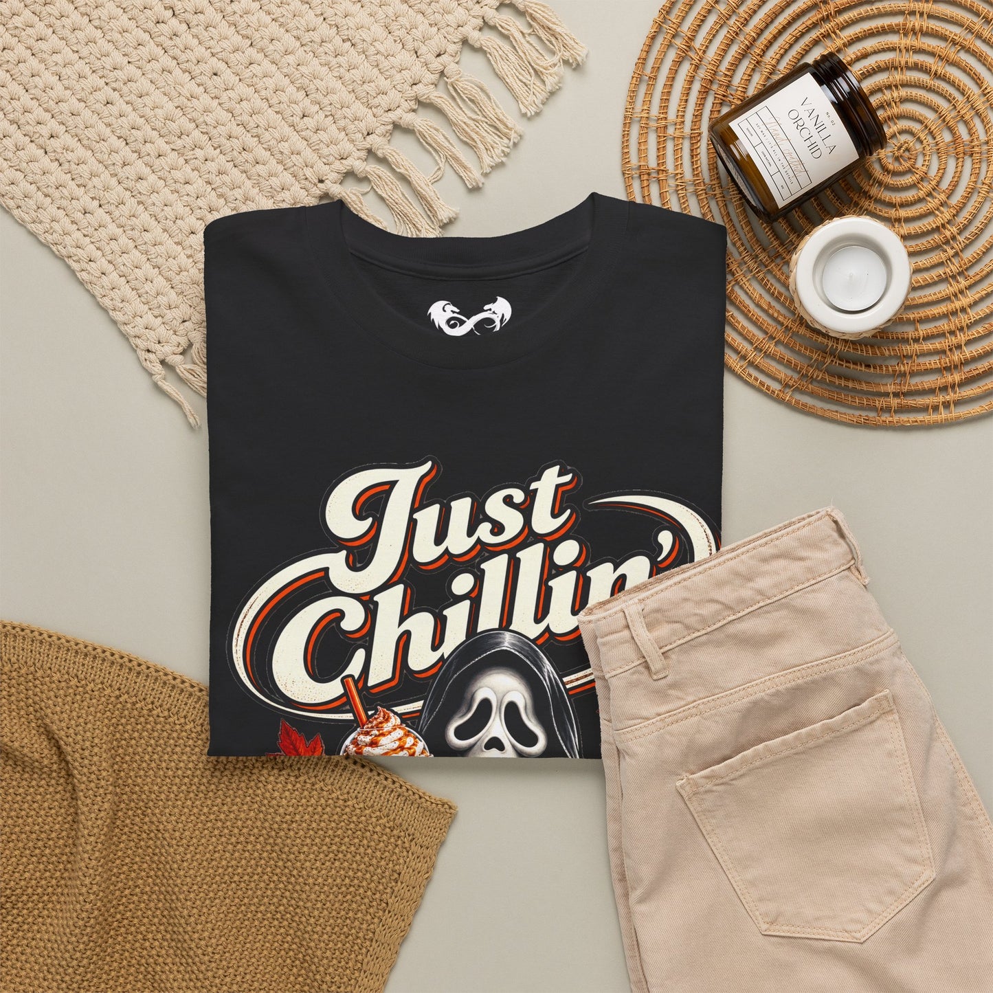 A folded black graphic T-shirt is styled in a flat lay with beige pants, a woven blanket, a candle jar, and a small cup on a neutral surface. The shirt features a large white and orange 'Just Chillin'' graphic with a Ghostface-style hooded figure and a small drink graphic near the bottom.
