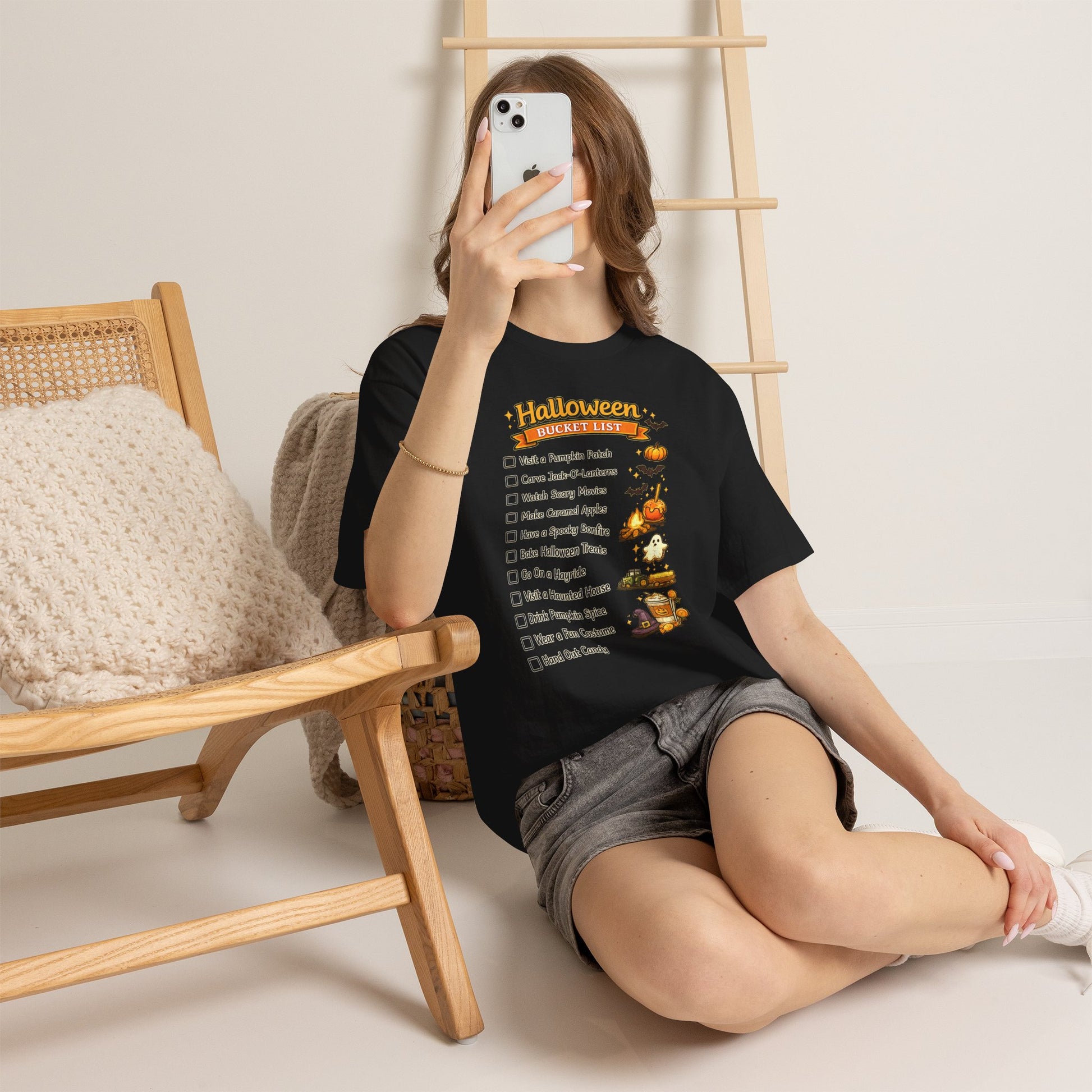 A woman sits on the floor in a minimalist room, taking a mirror selfie while wearing a black graphic t-shirt with a 'Halloween Chores List' design and gray denim shorts.