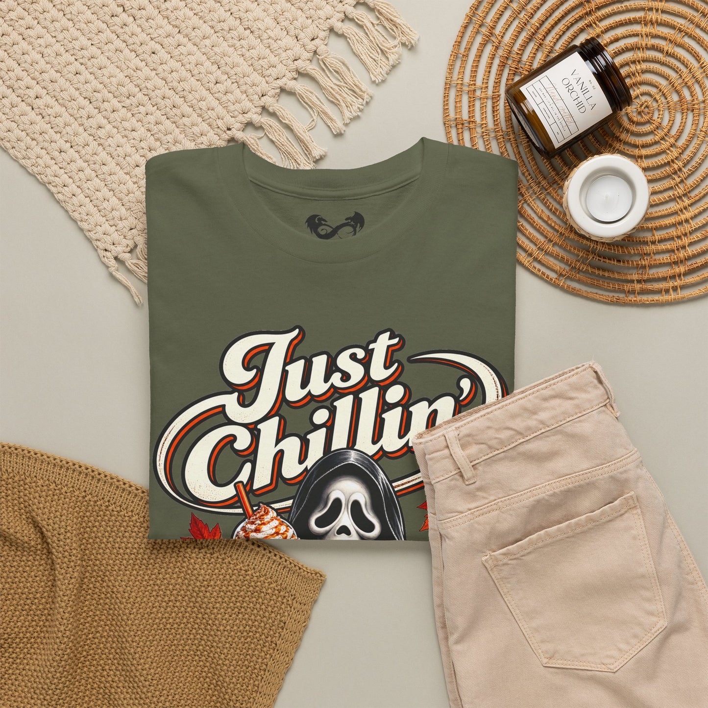 An olive green graphic t-shirt featuring the text 'Just Chillin'' in white with red and black outlines, alongside a Ghostface illustration, styled in a flat lay with beige pants, a knit blanket, and a candle.