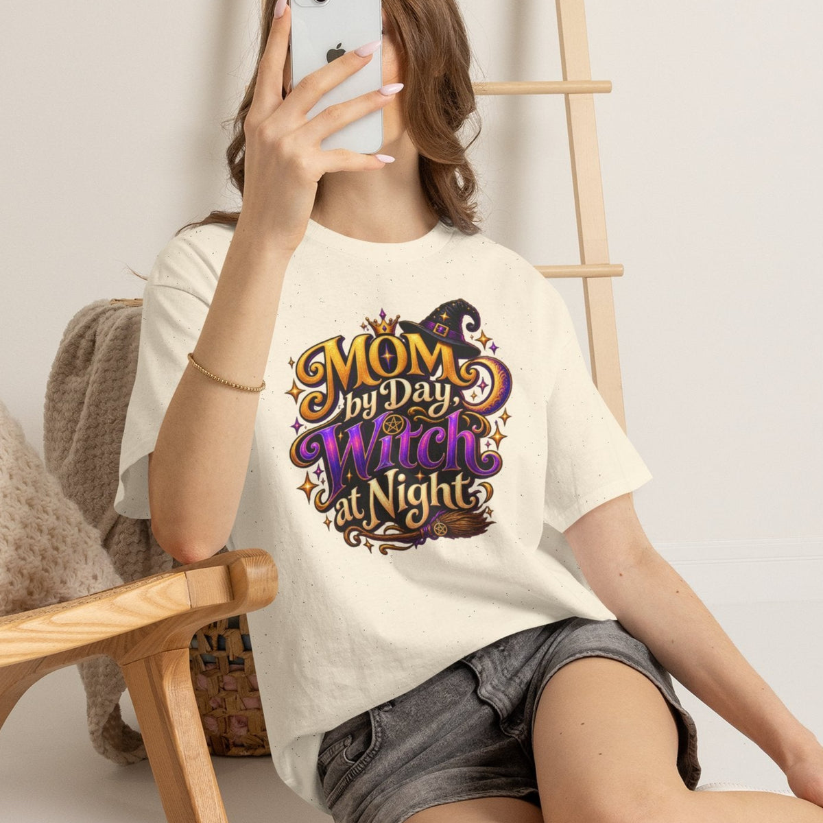 A woman takes a mirror selfie while wearing a cream-colored graphic t-shirt that reads 'MOM My Day My Witch Nights' in purple, gold, and black lettering. She is sitting on a light-colored floor next to a wooden chair with a knit pillow.