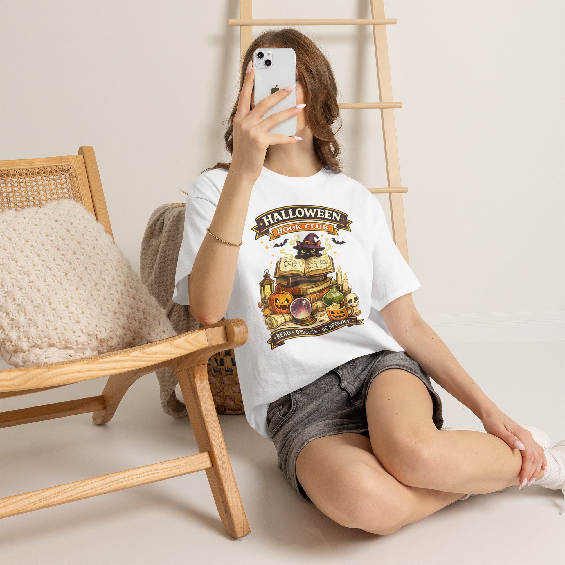 A woman takes a mirror selfie while wearing a white graphic t-shirt with a Halloween-themed treasure chest design and gray denim shorts, sitting on a light-colored floor next to a wooden chair.