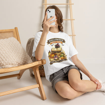 A woman takes a mirror selfie while wearing a white graphic t-shirt with a Halloween-themed treasure chest design and gray denim shorts, sitting on a light-colored floor next to a wooden chair.
