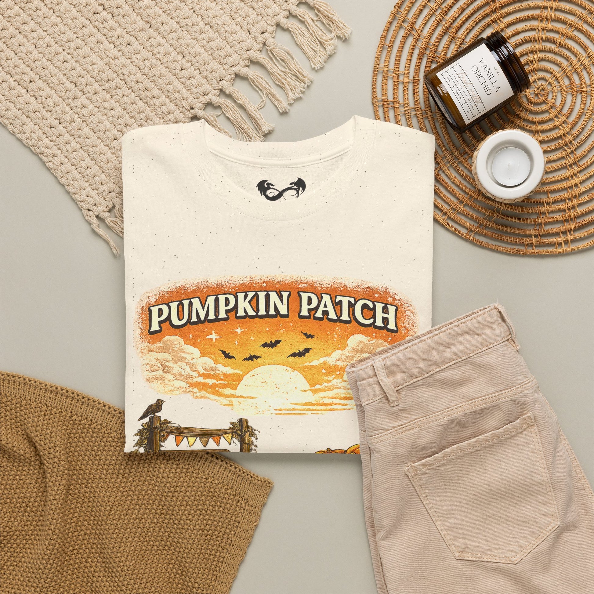 A folded cream graphic T-shirt with a Halloween-themed 'Pumpkin Patch' sunset print is styled in a flat lay with beige pants, a woven placemat, a candle jar, and a knit textile on a light gray surface.