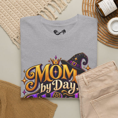 A folded heather gray t-shirt with a colorful 'MOM by Day' witch graphic, styled in a flat lay with beige pants, a woven blanket, a candle, and a cup on a neutral background.