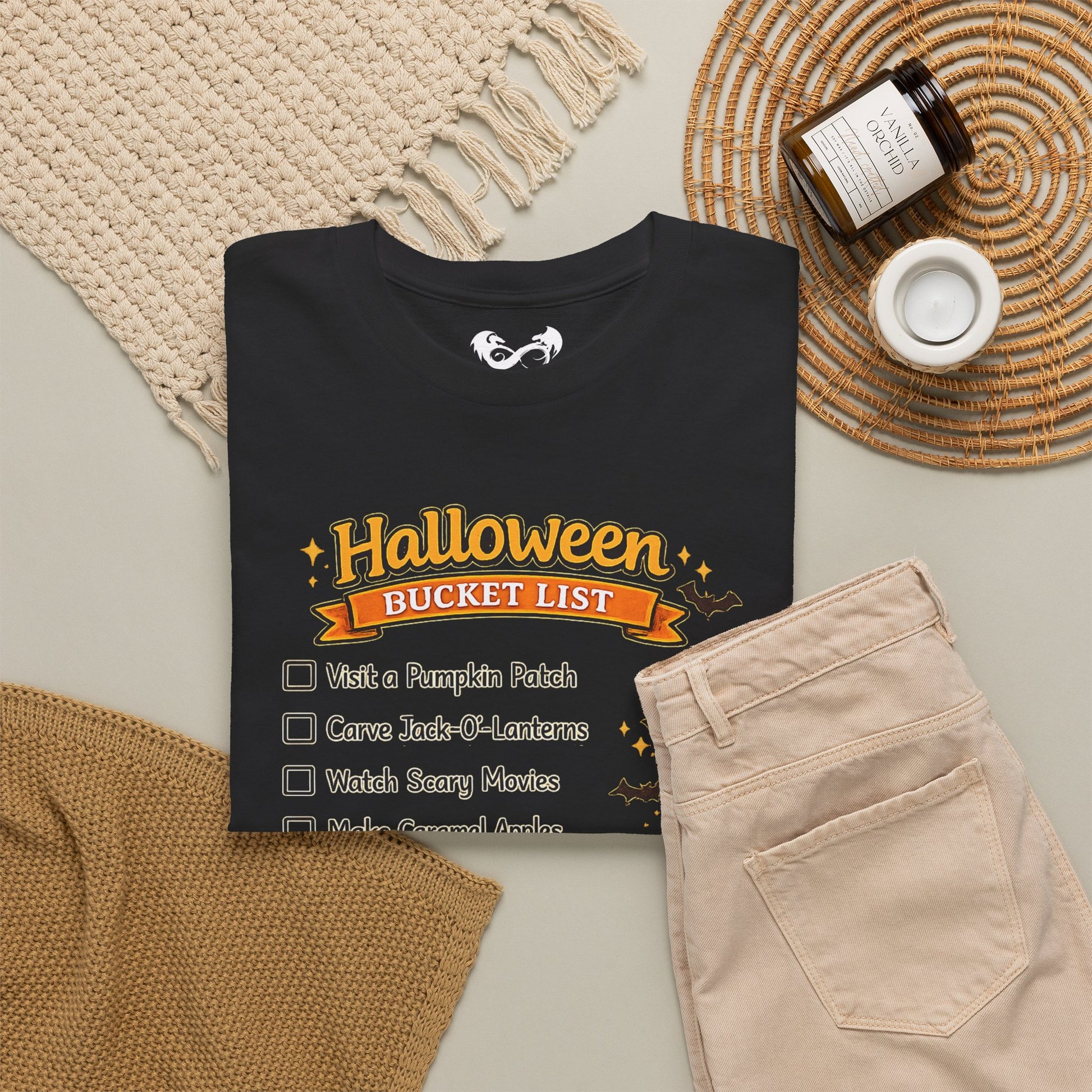 A folded black graphic T-shirt is arranged in a styled flat lay with beige pants, a woven blanket, a small candle jar, and a woven placemat on a light neutral background. The shirt graphic reads 'Halloween Bucket List' with checklist items below in orange, yellow, and white.