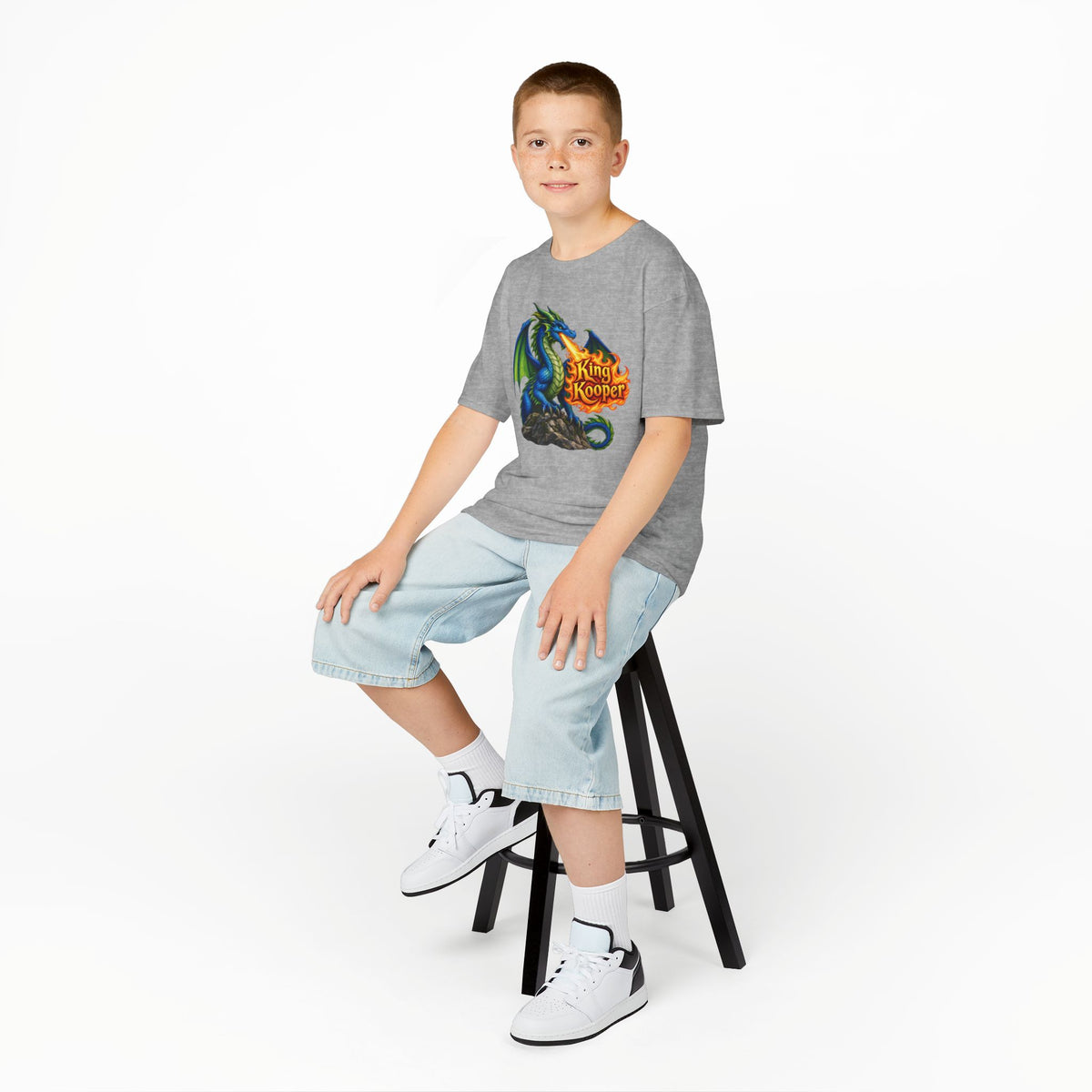 A boy sitting on a black stool wearing a heather gray t-shirt with a colorful graphic print, light wash denim shorts, white socks, and white sneakers, photographed against a seamless white studio background.