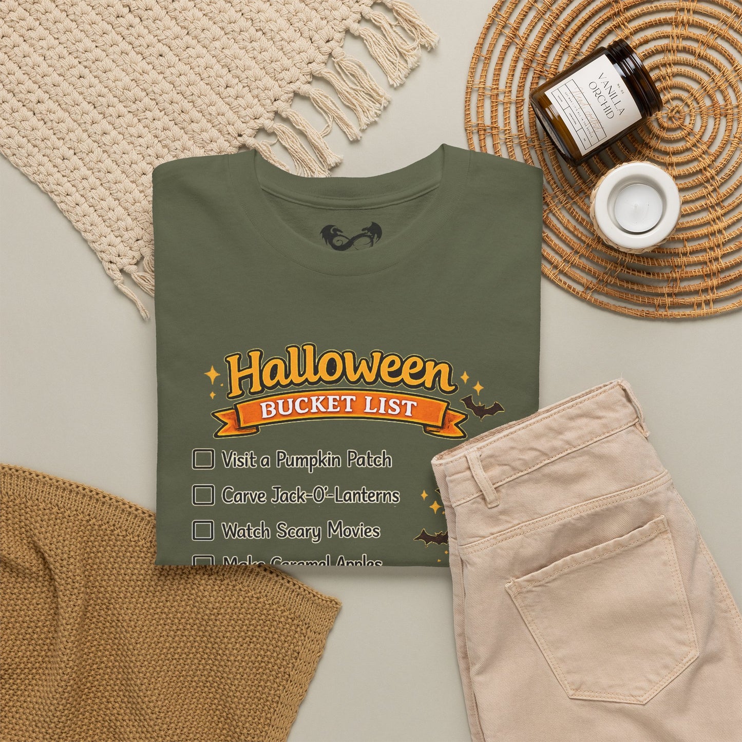 An olive green t-shirt with a 'Halloween Bucket List' graphic, styled in a flat lay with beige pants, a woven placemat, a knit blanket, and a candle.