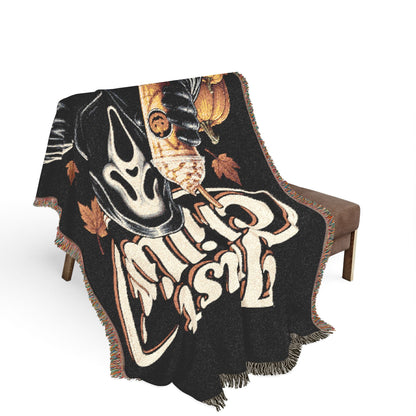 A black woven throw blanket with fringe, draped over a brown upholstered chair. The blanket features a large cream and orange graphic with a ghost face, a pumpkin, and stylized text reading 'Cult Leader'.