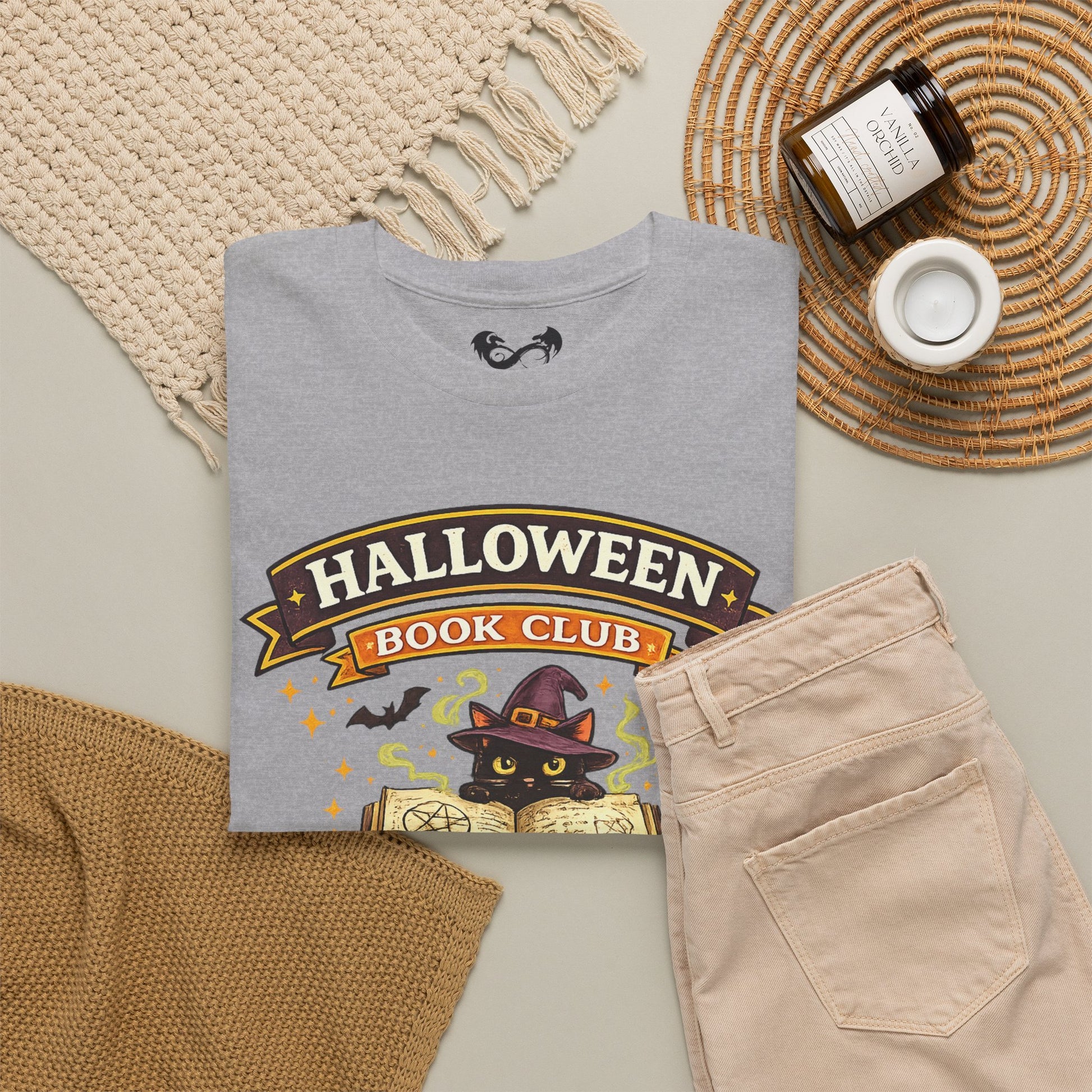 A folded light gray graphic T-shirt is styled in a flat lay with beige pants, a woven blanket, a candle jar, and a small cup on a neutral surface. The shirt graphic reads “HALLOWEEN BOOK CLUB” with a black cat in a witch hat and an open book.